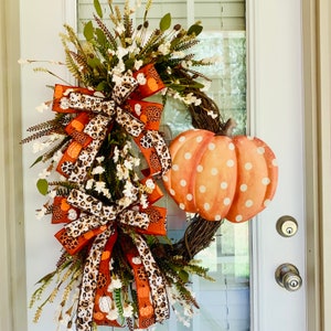 May include: A fall-themed wreath with orange and brown bows, white flowers, and a polka-dotted orange pumpkin. The wreath is decorated with small pumpkins and leaves.