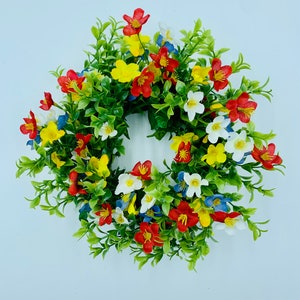 May include: A colorful floral wreath with red, yellow, white, and blue flowers. The wreath is made of artificial flowers and greenery.