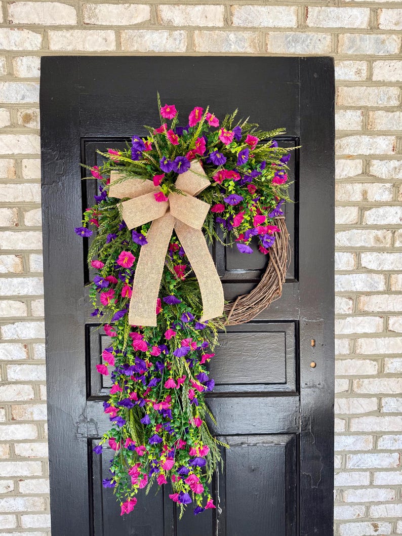 May include: A vibrant floral wreath with pink and purple flowers, green foliage, and a tan burlap bow. The wreath is displayed on a black door against a brick wall, creating a welcoming aesthetic.