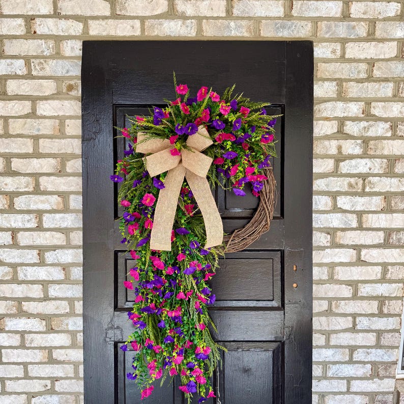 May include: A vibrant floral wreath with pink and purple flowers, green foliage, and a tan burlap bow. The wreath is displayed on a black door against a brick wall, creating a welcoming entrance.