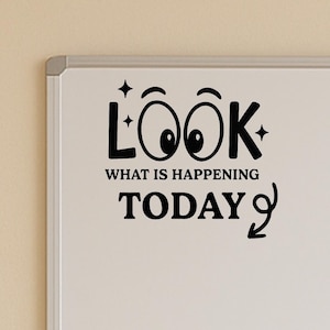 May include: A white dry-erase board with black text that reads "LOOK" with stylized eyes, "WHAT IS HAPPENING", and "TODAY". The board has a silver frame and is mounted on a beige wall.