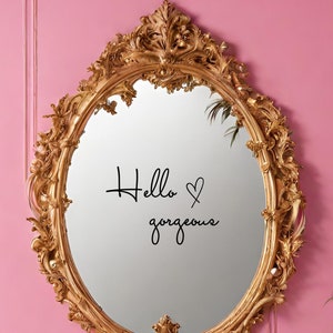 May include: An ornate, gold-colored oval mirror with intricate detailing. The mirror's surface reflects the words "Hello gorgeous" in black script. The mirror is set against a pink wall, creating a decorative accent.