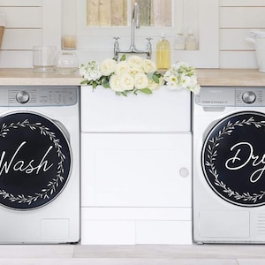 May include: Two white washing machines with black and white floral wreath decals. The left machine has the word "Wash" in white lettering on the decal. The right machine has the word "Dry" in white lettering on the decal.