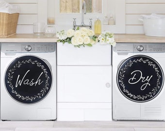 May include: Two white washing machines with black and white floral wreath decals. The left machine has the word "Wash" in white lettering on the decal. The right machine has the word "Dry" in white lettering on the decal.