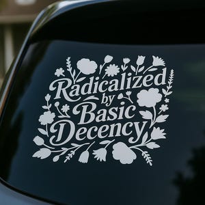 May include: White vinyl decal on a car window. The decal features the text "Radicalized by Basic Decency" in a decorative font, surrounded by floral and leaf illustrations. The car window is black.