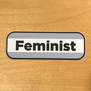 Può includere: Un adesivo rettangolare grigio e bianco con angoli arrotondati e un bordo nero. L'adesivo dice "Femminista" in testo nero.