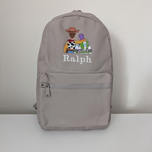 May include: A light grey backpack featuring an embroidered graphic of two cartoon characters sitting side-by-side with arms around each other. Below the embroidery, the name Ralph is stitched in white thread. The bag has a front zippered pocket.