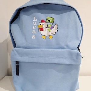 May include: A light blue backpack featuring an embroidered design of a green pixelated character riding a white chicken. The name Lucas is embroidered vertically in white letters to the left of the graphic. The bag has a front zippered pocket.