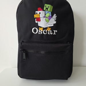 May include: A black backpack featuring an embroidered design of a green block-style character riding a white block-style chicken. Below the embroidery, the name Oscar is stitched in white lettering. The bag includes a front zippered pocket.