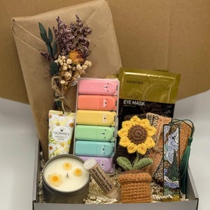 May include: A gift box filled with assorted items. Includes a candle, lip balm, eye mask, highlighters, a crochet sunflower, and a bookmark. The box is lined with paper and has a brown paper-wrapped gift and dried flowers.