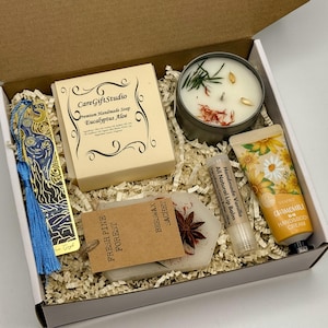 May include: An open gift box filled with a variety of items. Includes a candle, soap, lip balm, hand cream, a bookmark, and a sachet. The box contains the text "CareGiftStudio" and "Fresh Pine Forest". The items are arranged on shredded paper.