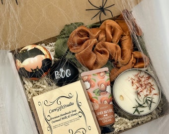 May include: A Halloween-themed gift box filled with a variety of items. Includes a black and orange bat-shaped item, a black item with "BOO" written on it, a bar of soap, a tube of shea butter hand cream, a candle, and scrunchies. The box is decorated with spiderwebs and spiders.