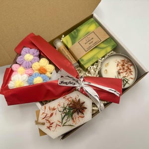 May include: A gift box containing a bar of green lemon verbena soap, a vanilla lip balm, a round candle with dried petals, and a white wax melt decorated with star anise and dried herbs, plus a bouquet of small crocheted flowers in a red wrap.