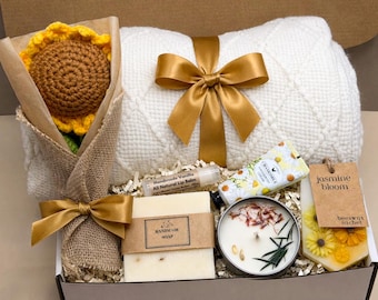 May include: A gift box filled with a crochet sunflower bouquet, a white textured blanket tied with a gold bow, handmade soap, lip balm, chamomile cream, a candle, and a jasmine bloom sachet. The box is lined with shredded paper.