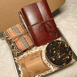 May include: A gift box filled with a brown leather journal, a pen, a knitted scarf, a small brown bag with the text "10 Tea Infuser Bags" and the word "Enjoy", and a small tin of loose leaf tea.