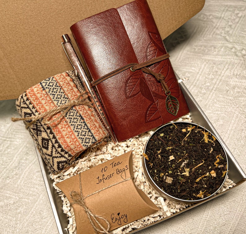 May include: A gift box filled with a brown leather journal, a pen, a knitted scarf, a small brown bag with the text "10 Tea Infuser Bags" and the word "Enjoy", and a small tin of loose leaf tea.