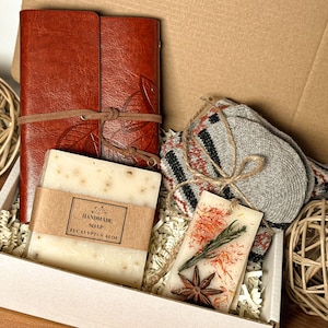 May include: A white gift box filled with a brown leather journal, a bar of handmade eucalyptus aloe soap, a pair of gray knitted socks, and a small candle with dried flowers and star anise.