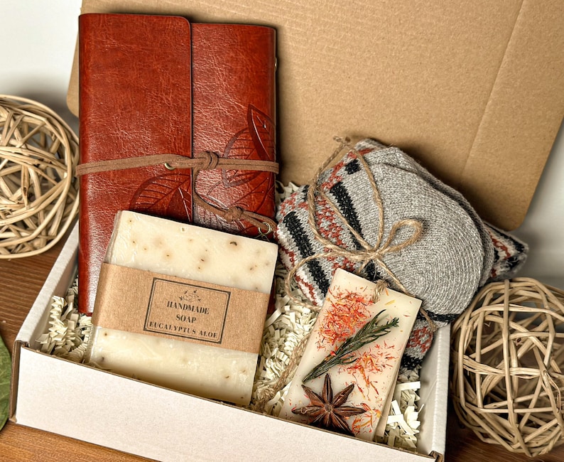 May include: A white gift box filled with a brown leather journal, a bar of handmade eucalyptus aloe soap, a pair of gray knitted socks, and a small candle with dried flowers and star anise.