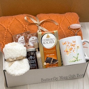 May include: A gift box containing an orange knit blanket, a white mug with butterfly and dandelion graphics, a Ferrero Rocher chocolate bar, a jar of honey, two tea bags, a lip balm, and a pink facial roller. The box is labelled Butterfly Mug Gift Box.