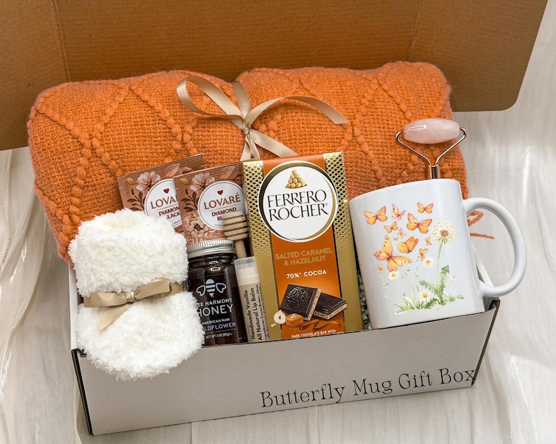May include: A gift box containing an orange knit blanket, a white mug with butterfly and dandelion graphics, a Ferrero Rocher chocolate bar, a jar of honey, two tea bags, a lip balm, and a pink facial roller. The box is labelled Butterfly Mug Gift Box.