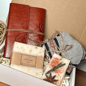 May include: A gift box filled with a brown leather journal, a bar of handmade eucalyptus aloe soap, a gray and red striped knitted scarf, and a small candle with dried flowers and star anise.
