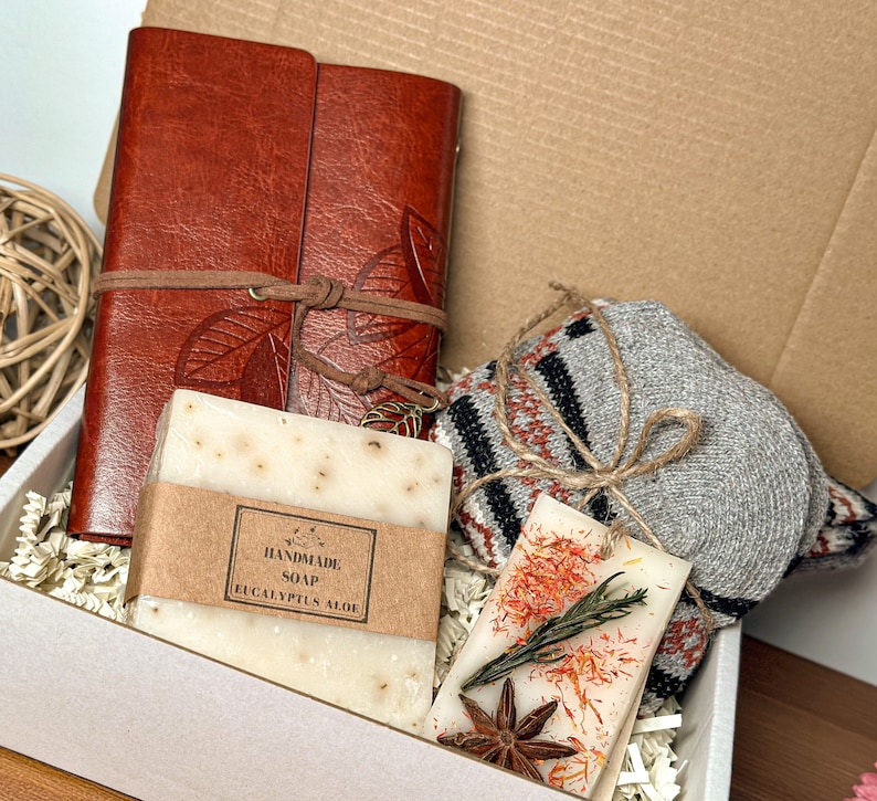 May include: A gift box filled with a brown leather journal, a bar of handmade eucalyptus aloe soap, a gray and red striped knitted scarf, and a small candle with dried flowers and star anise.