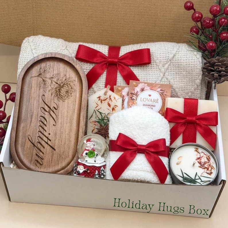 May include: A gift box labelled Holiday Hugs Box containing a wooden tray engraved with the name Kaitlyn, a white knit blanket with a red ribbon, a small snow globe, a white candle, a bar of soap with a red ribbon, and tea packets.