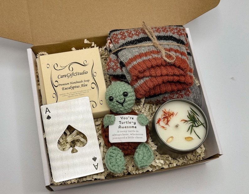 May include: A gift box containing a bar of soap, a metal Ace of Spades card, a crocheted turtle, a candle, and a pair of socks. The soap is labeled "CareGiftStudio Eucalyptus Aloe." The turtle has a tag that reads "You're Turtle-y Awesome."