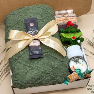 May include: A winter-themed gift box with a green blanket tied with a gold ribbon. The box contains a cappuccino packet, a small crocheted Christmas tree, a candle, lip balm, and wrapped candies.