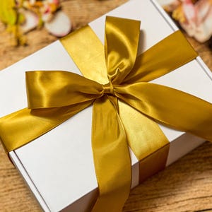 May include: A rectangular white cardboard gift box wrapped with a wide, shiny gold satin ribbon tied in a large, centered bow. The ribbon crosses the top of the box horizontally and vertically, creating a clean, elegant presentation for the container.