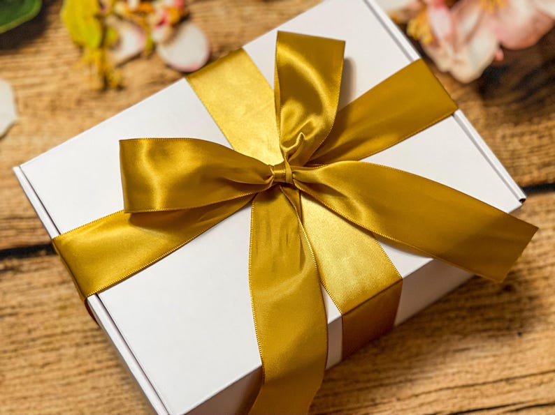 May include: A rectangular white cardboard gift box wrapped with a wide, shiny gold satin ribbon tied in a large, centered bow. The ribbon crosses the top of the box horizontally and vertically, creating a clean, elegant presentation for the container.