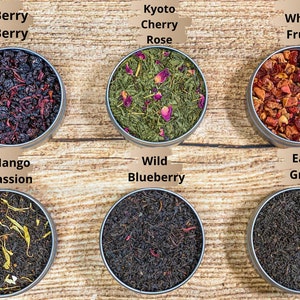 Op de afbeelding: Zes ronde metalen blikken met losse bladthee, elk met een andere mix. De blikken zijn gerangschikt in twee rijen van drie. De bovenste rij van links naar rechts is Berry Berry, Kyoto Cherry Rose en Whole Fruits. De onderste rij van links naar rechts is Mango Passion, Wild Blueberry en Earl Grey.