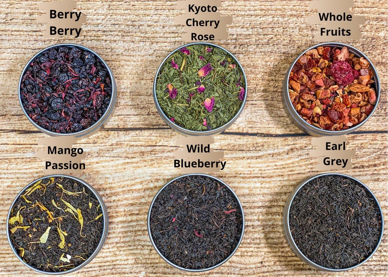 Op de afbeelding: Zes ronde metalen blikken met losse bladthee, elk met een andere mix. De blikken zijn gerangschikt in twee rijen van drie. De bovenste rij van links naar rechts is Berry Berry, Kyoto Cherry Rose en Whole Fruits. De onderste rij van links naar rechts is Mango Passion, Wild Blueberry en Earl Grey.
