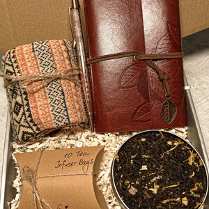 May include: A gift box filled with a brown leather journal, a pen, a pair of socks, a brown paper bag with the text "10 Tea Infuser Bags" and the word "Enjoy", and a tin of loose leaf tea.