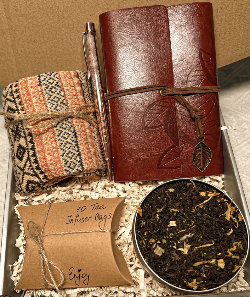 May include: A gift box filled with a brown leather journal, a pen, a pair of socks, a brown paper bag with the text "10 Tea Infuser Bags" and the word "Enjoy", and a tin of loose leaf tea.