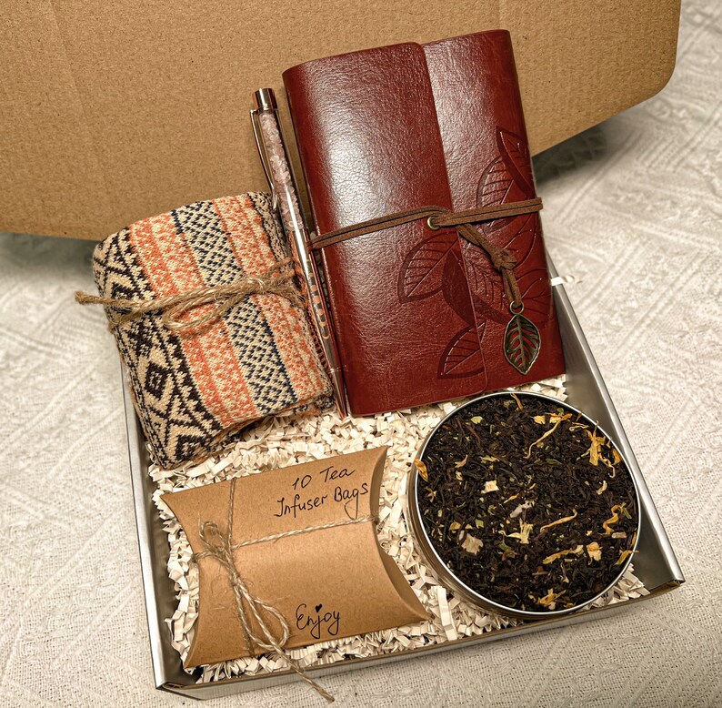 May include: A gift box filled with a brown leather journal with a leaf design, a brown and orange striped knitted scarf, a brown paper bag with the text "10 Tea Infuser Bags" and the word "Enjoy", and a small tin of loose leaf tea.