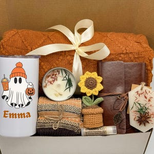 May include: A gift box filled with a white tumbler featuring a ghost illustration, a candle, a brown leather journal, a crocheted sunflower, a lip balm, and a cozy orange blanket tied with a cream ribbon.