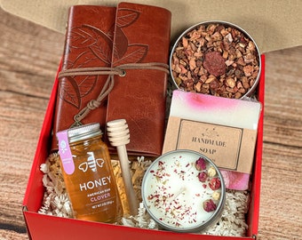 May include: A red gift box filled with a variety of items, including a brown leather journal, a jar of honey, a wooden honey dipper, a candle with dried rose petals, a bar of soap, and a tin of loose leaf tea.