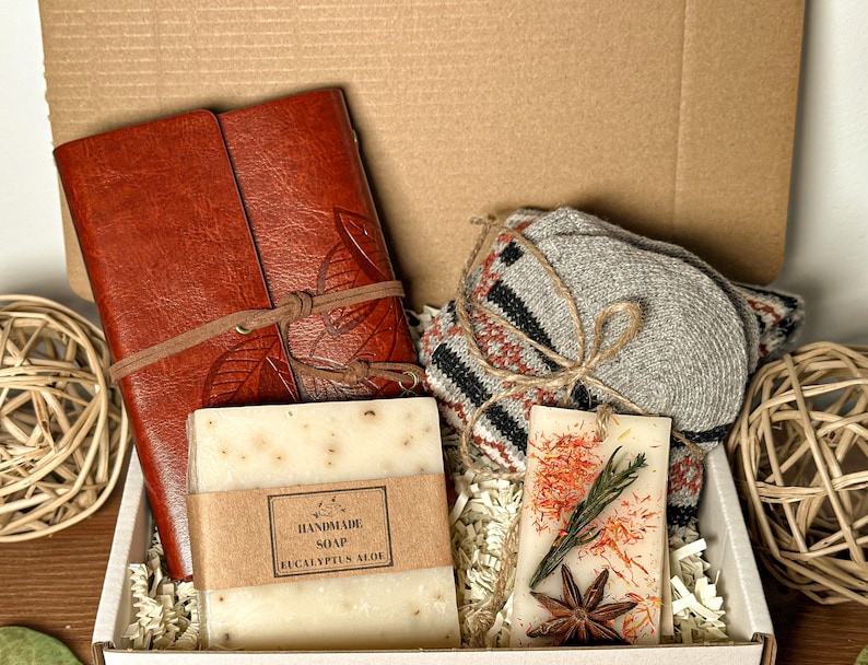 May include: A gift box filled with a brown leather journal, a bar of handmade eucalyptus aloe soap, a scented candle with dried flowers and star anise, and a pair of gray knitted socks.