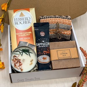 May include: A gift box containing a Ferrero Rocher Salted Caramel & Hazelnut chocolate bar, a Taste instant cappuccino packet, a folded orange and brown patterned knit item, a round candle with botanical toppings, and a bar of eucalyptus aloe soap.