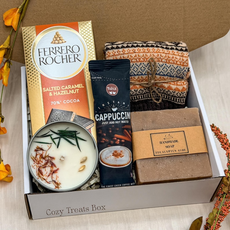 May include: A gift box containing a Ferrero Rocher Salted Caramel & Hazelnut chocolate bar, a Taste instant cappuccino packet, a folded orange and brown patterned knit item, a round candle with botanical toppings, and a bar of eucalyptus aloe soap.