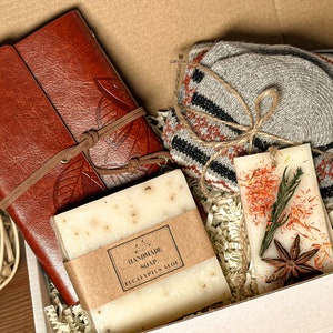 May include: A gift box filled with a brown leather journal, a bar of handmade eucalyptus soap, a scented candle with dried flowers and star anise, and a pair of gray knitted socks.