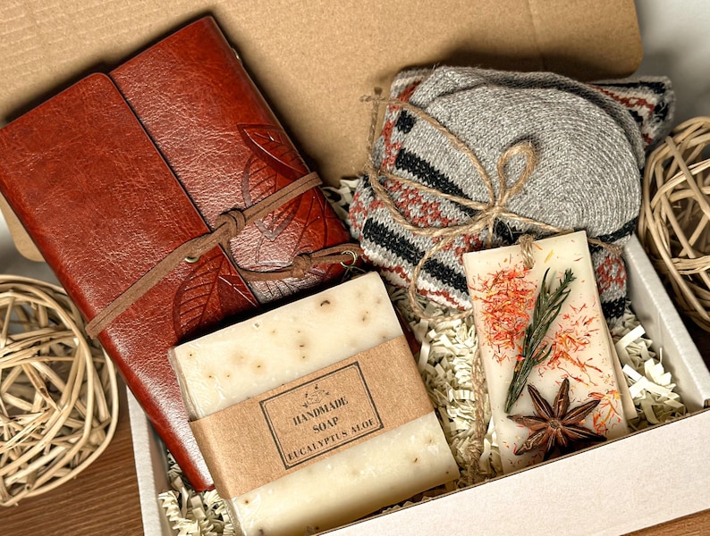 May include: A gift box filled with a brown leather journal, a bar of handmade eucalyptus soap, a scented candle with dried flowers and star anise, and a pair of gray knitted socks.