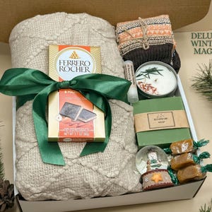 May include: A gift box containing a beige knit blanket tied with a green ribbon, a Ferrero Rocher chocolate bar, a pair of patterned socks, a candle, a lip balm, a bar of handmade soap, a small snow globe, and three wrapped caramels.