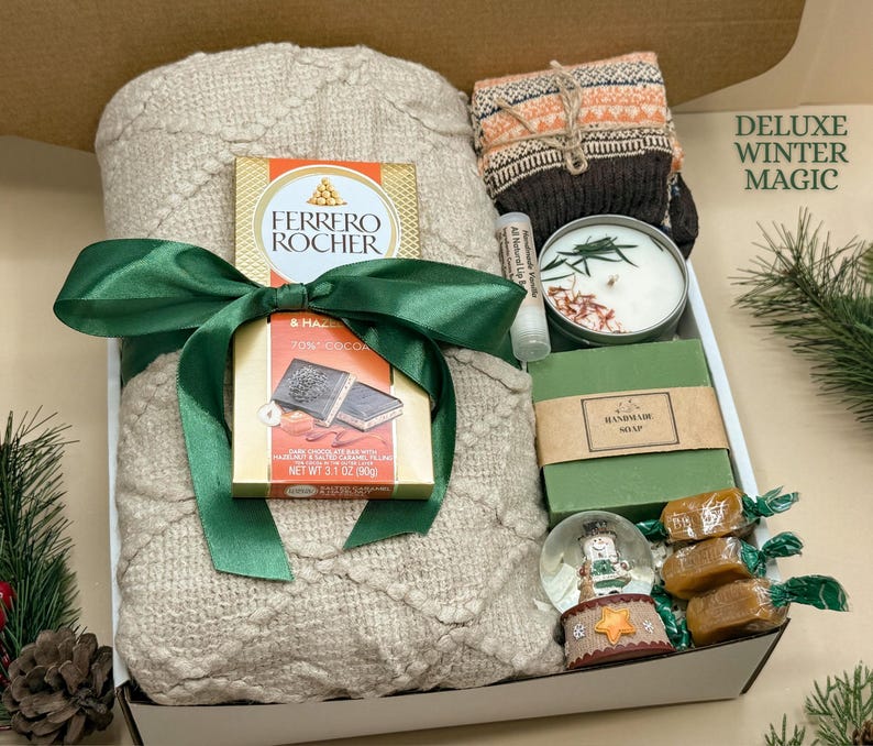 May include: A gift box containing a beige knit blanket tied with a green ribbon, a Ferrero Rocher chocolate bar, a pair of patterned socks, a candle, a lip balm, a bar of handmade soap, a small snow globe, and three wrapped caramels.