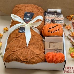 May include: A gift box containing a burnt orange textured knit blanket tied with a cream ribbon, a ceramic orange pumpkin, a bar of handmade soap, a pair of patterned socks, a vanilla lip balm, and a small crocheted orange pumpkin keychain.