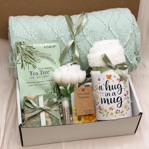 May include: A gift box containing a light green textured throw blanket, a Tea Tree collagen essence mask, a white flower-shaped candle, a green marbled soap, a lip balm, a jasmine bloom beeswax sachet, and a white mug labeled a hug in a mug.