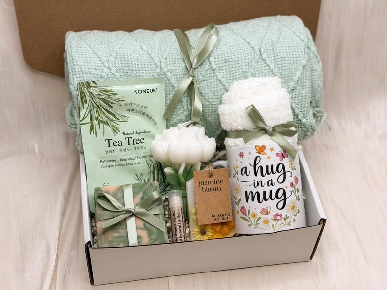 May include: A gift box containing a light green textured throw blanket, a Tea Tree collagen essence mask, a white flower-shaped candle, a green marbled soap, a lip balm, a jasmine bloom beeswax sachet, and a white mug labeled a hug in a mug.