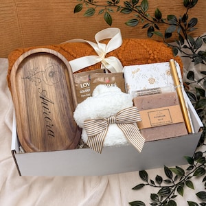 May include: A gift box containing an oval wooden tray engraved with a floral design and the name Aurora, a white fuzzy item with a striped bow, a bar of handmade soap, a gold pen, a notebook, and tea packets, all resting on a burnt orange knit blanket.