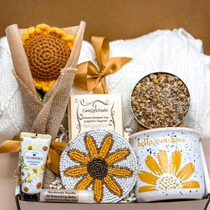 May include: A gift box containing a crocheted sunflower in burlap, a white cable-knit blanket with a gold ribbon, a CareGiftStudio soap, a tin of dried chamomile, a beaded sunflower coaster, a chamomile hand cream, and a hello sunshine mug.
